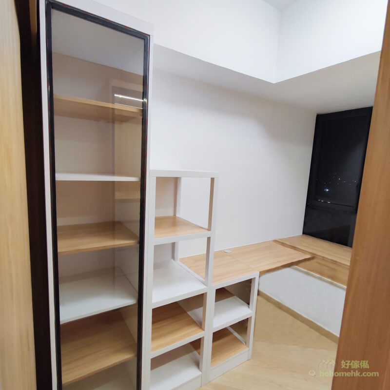 角落衣帽間一隅，左邊深啡邊框玻璃門高身衣櫃內有原木色層板，中間白色框配淺木層板的開放式階梯層架，右後方接駁原木色地台坐墊，落地深色窗外見到城市夜景。