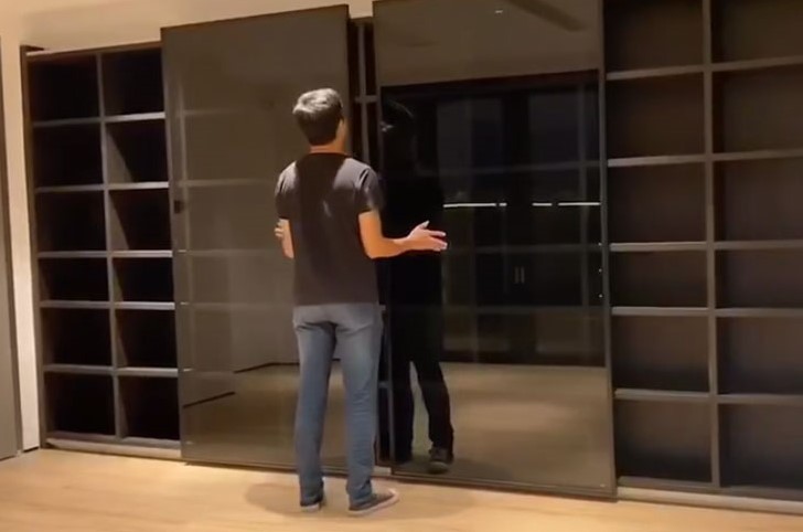 一名穿深灰短袖男士背向鏡頭，喺空置單位內單手推動一道深色茶玻璃懸吊式趟門，左右兩邊係深木色開放式格仔櫃，地面鋪淺色木地板，玻璃門滑至中間位置，示範懸吊滑軌嘅順滑推拉效果。