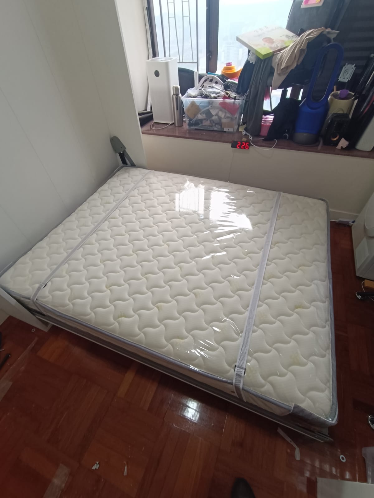 從上方斜拍已打開嘅白色Livo直翻變形床，白色波浪紋床褥外包透明膠套，兩條灰色固定帶由床頭伸向床尾，床身置於窗台旁嘅木地板上。