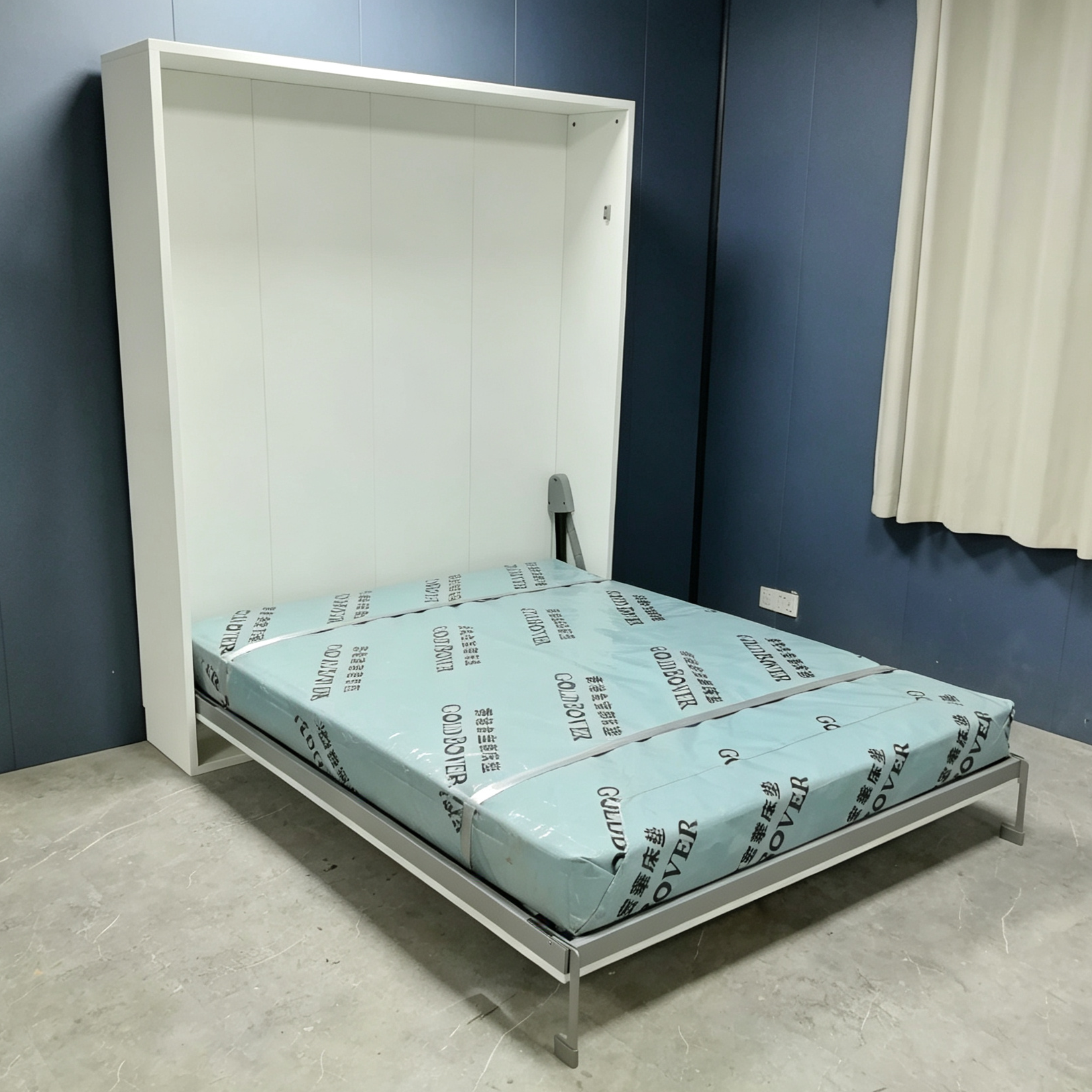 右前方斜角拍攝打開後嘅白色Livo直翻變形床，淺藍保護套床褥由灰色帶扣穩，床褥放於淺灰金屬床框上，白色高身櫃靠住深藍牆身。