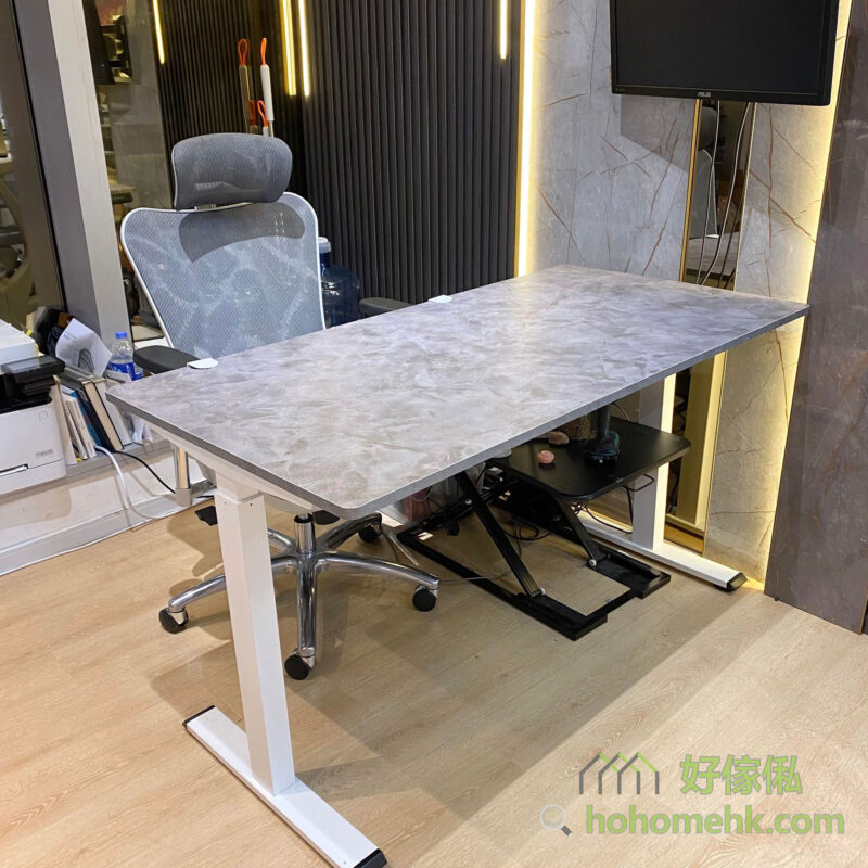 展銷廳實物照，Morton電動升降枱正面全景，呈現水泥灰仿石紋枱面配白色T形電動升降鋼架，枱下可見黑色折疊踏腳架，灰色辦公椅置於枱後，暖黃燈帶牆面背景，右側有ASUS電腦顯示器，木紋地板。