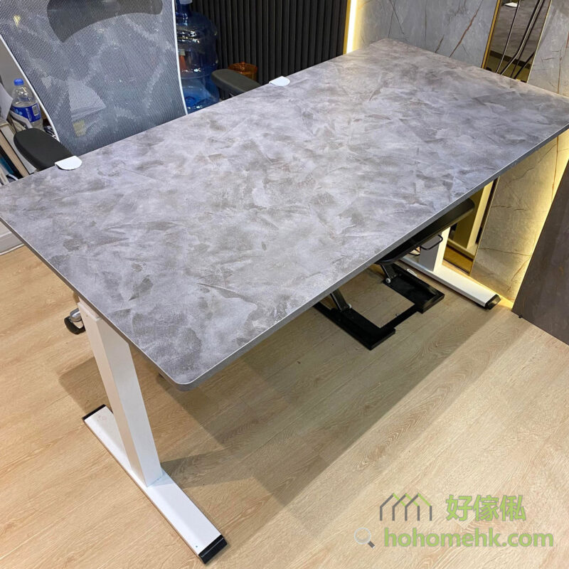 展銷廳實物照，Morton電動升降枱呈現水泥灰仿石紋枱面配白色T形電動升降鋼架，枱面正上方俯視拍攝，可見圓角設計及紋理細節，四角附有白色防撞護角，木紋地板，背景有灰色辦公椅。