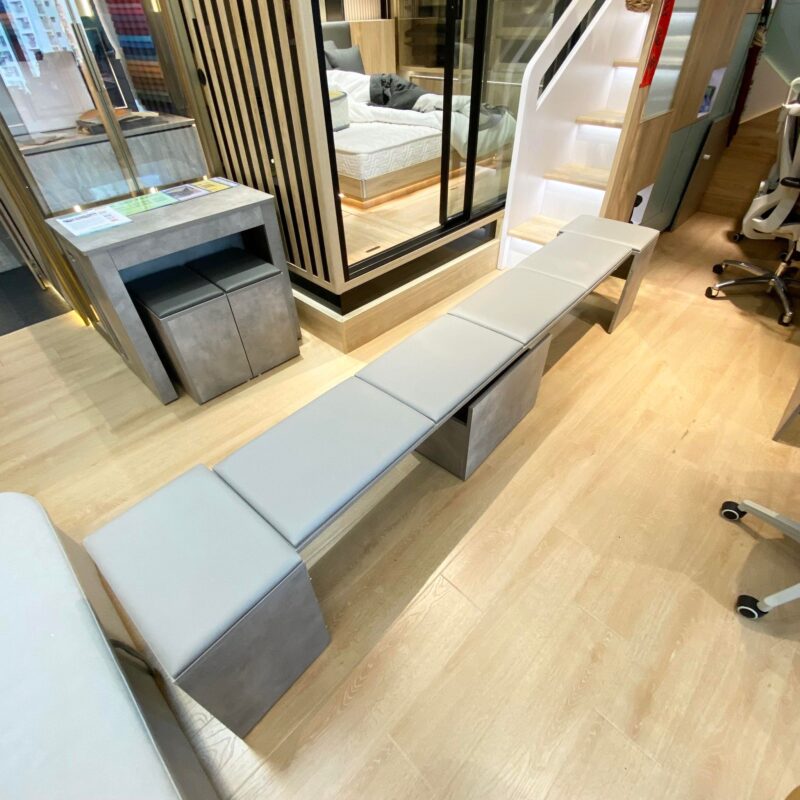 Vito+ 5人伸縮椅D8210布魯灰石色款門市實拍，椅子完全拉開成長凳，淺灰坐墊由一端延伸至另一端，椅身深灰水泥色，背景門市內可見同款縮起狀態及其他展示枱，木色地板，左方有玻璃幕牆及其他系統櫃。