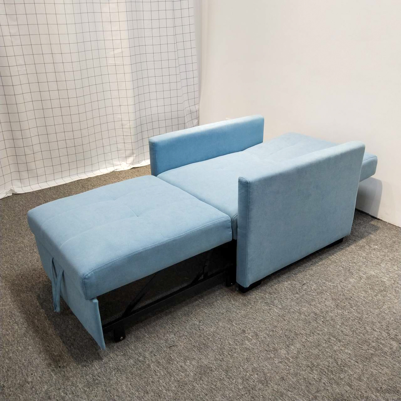 Cerulean 7279款梳化床完全展開為床鋪模式，靠背幾近放平，延伸座台完全拉出形成長方形睡眠面，整體淺藍色絨布面料鋪滿，右方扶手豎立，底部黑色金屬支架支撐，從右後方斜角拍攝，背景為白色格仔布幕及灰色地氈。