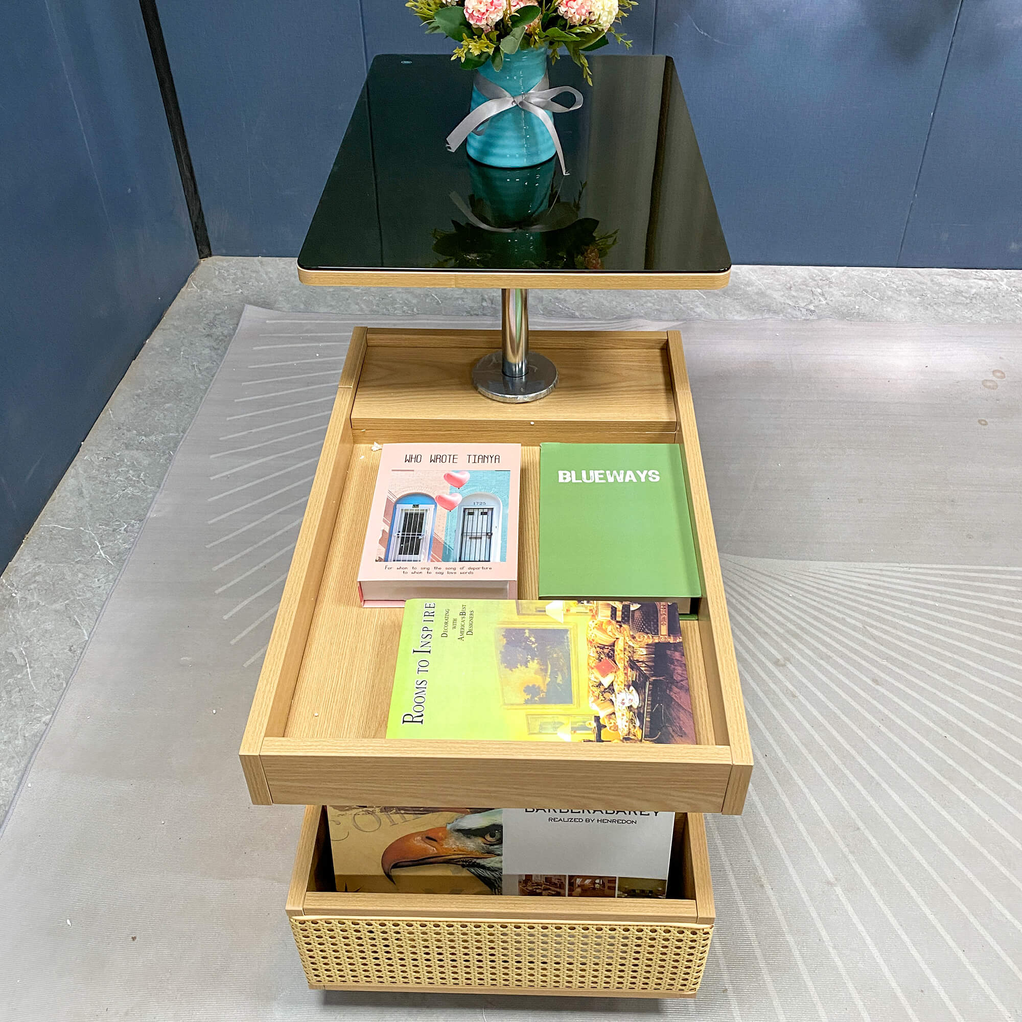 Parker升降茶几俯視圖，旋轉展開狀態，黑色玻璃枱面旋至一角可見花瓶反影，原木色上層開放托盤放有粉色封面、綠色封面及黃色封面書本各一，下層籐編籃放有大型藝術書冊，整體以原木色為主調，背景為淺灰色地板。