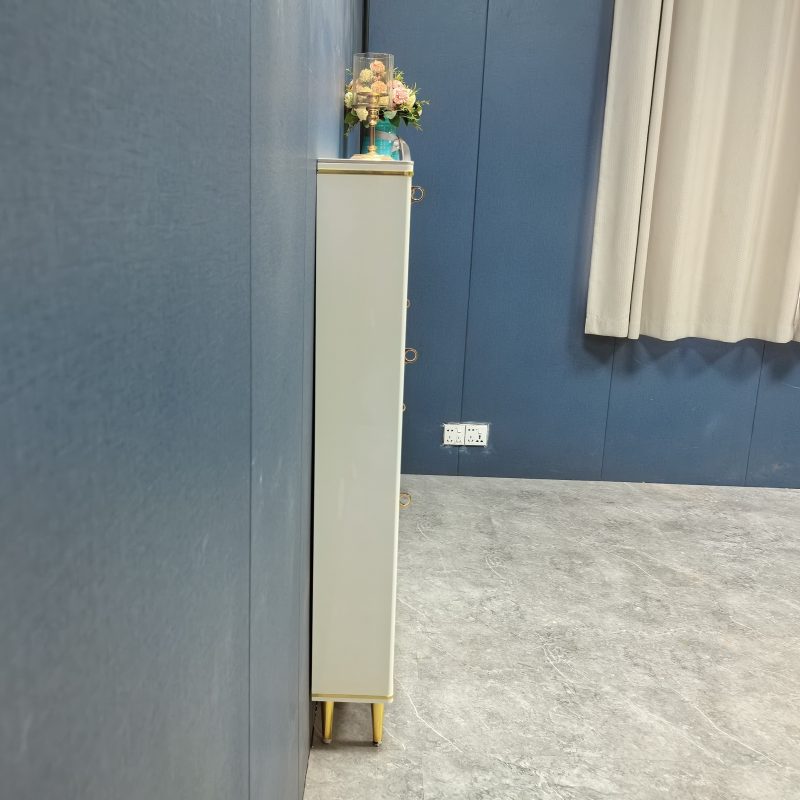 米白色亮光焗漆鞋櫃九十度側面實拍，顯示櫃身極薄輪廓，櫃頂擺放金色高腳玻璃燭台同粉色小花藝，右邊掛淺色窗簾，牆上有白色雙位電源插座，地面為灰色雲石紋磚，牆身為深藍色布紋。