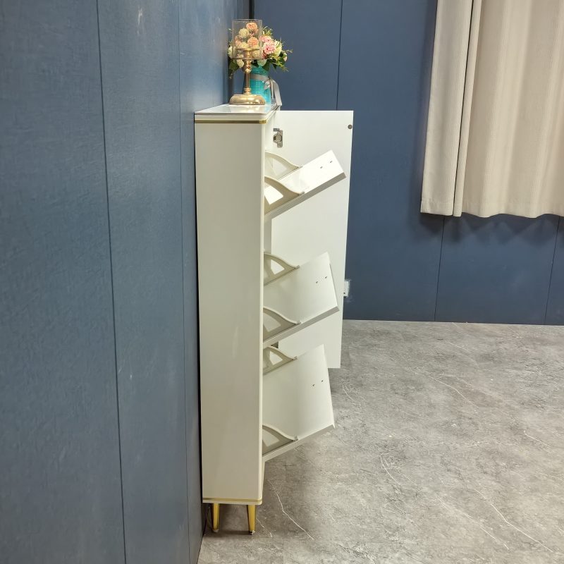 Caleb 超薄鞋櫃工廠側面實拍，深藍色牆前米白櫃身三個翻斗打開，露出向前斜放嘅白色層板，櫃頂擺粉紅花球同金色檯燈，地面為灰紋雲石。