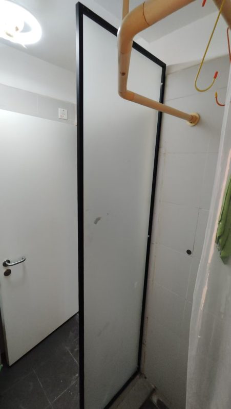 浴室入面近距離影黑色鋁框配磨砂玻璃單片浴屏，由地下豎到天花，左邊係白色廁所門，右上角望到銅色喉管同毛巾架，地下鋪深灰色方形磁磚。